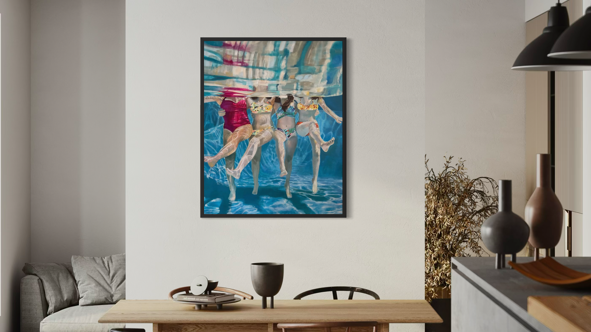 A framed colorful painting of three women in swimwear hanging on a white wall in a modern dining room with a wooden table, chairs, and pendant lights.